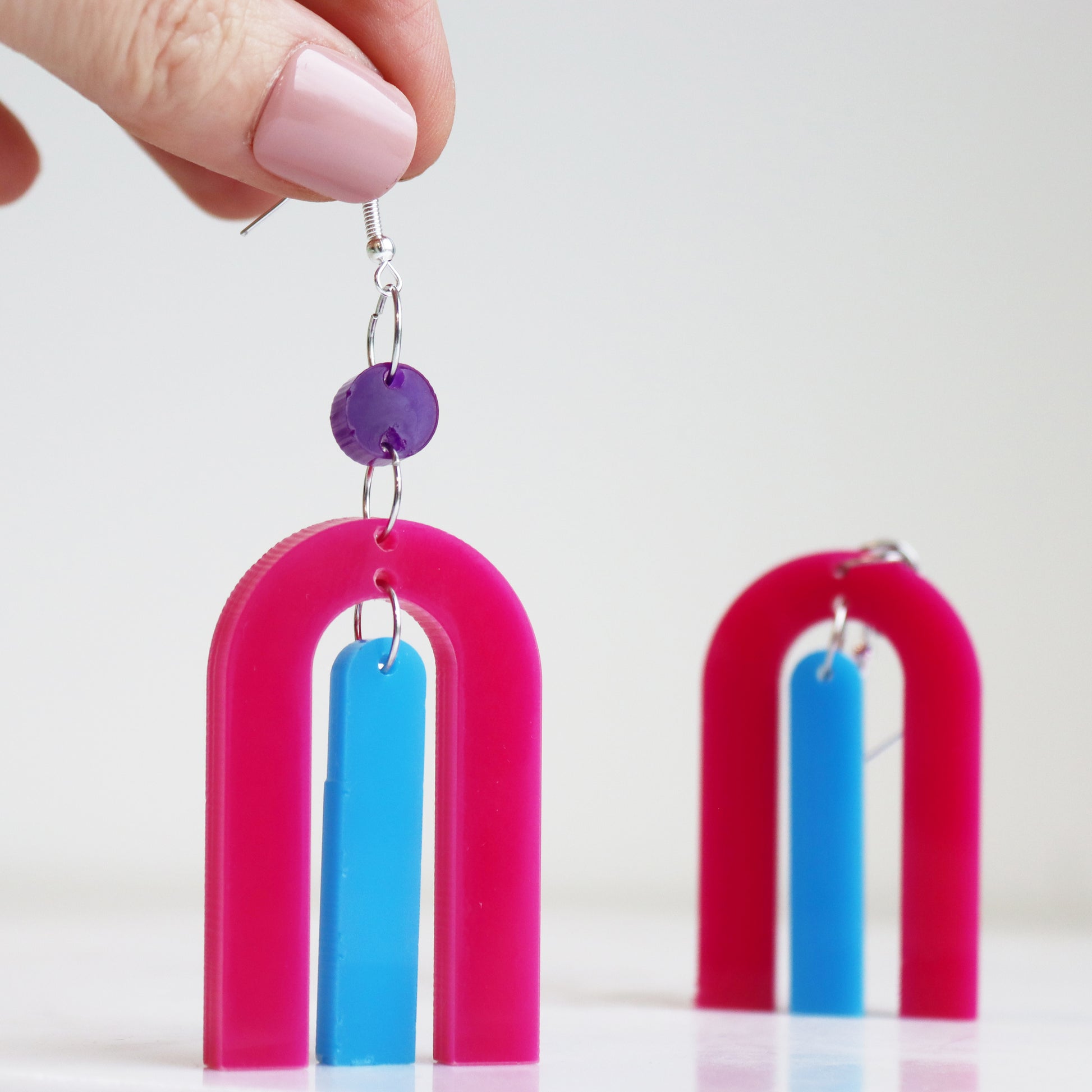 modern bright and colourful geometric arch dangle earrings cut from a purple, pink and turquoise acrylic shown hanging from a hand