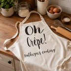 Personalised 'Oh Crepe!' apron with 'Kathryn's cooking' on a wooden kitchen worktop
