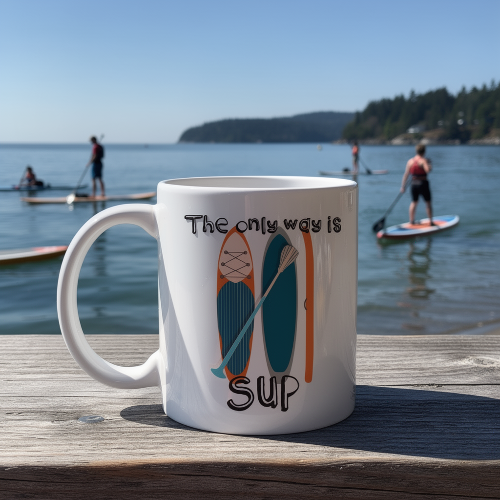 White mug with paddleboard design and 'The only way is SUP' text on a wooden dock with people paddleboarding in the background.