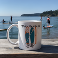 White mug with paddleboard design and 'The only way is SUP' text on a wooden dock with people paddleboarding in the background.