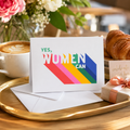 'Yes, Women Can' card with bold rainbow lettering, beside a coffee and flowers