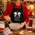 Black 'We whisk you a merry Christmas' apron with whisk and bow, worn while baking in a festive kitchen