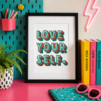 Framed 'Love Yourself' motivational print in retro teal and red lettering on a bright pink desk