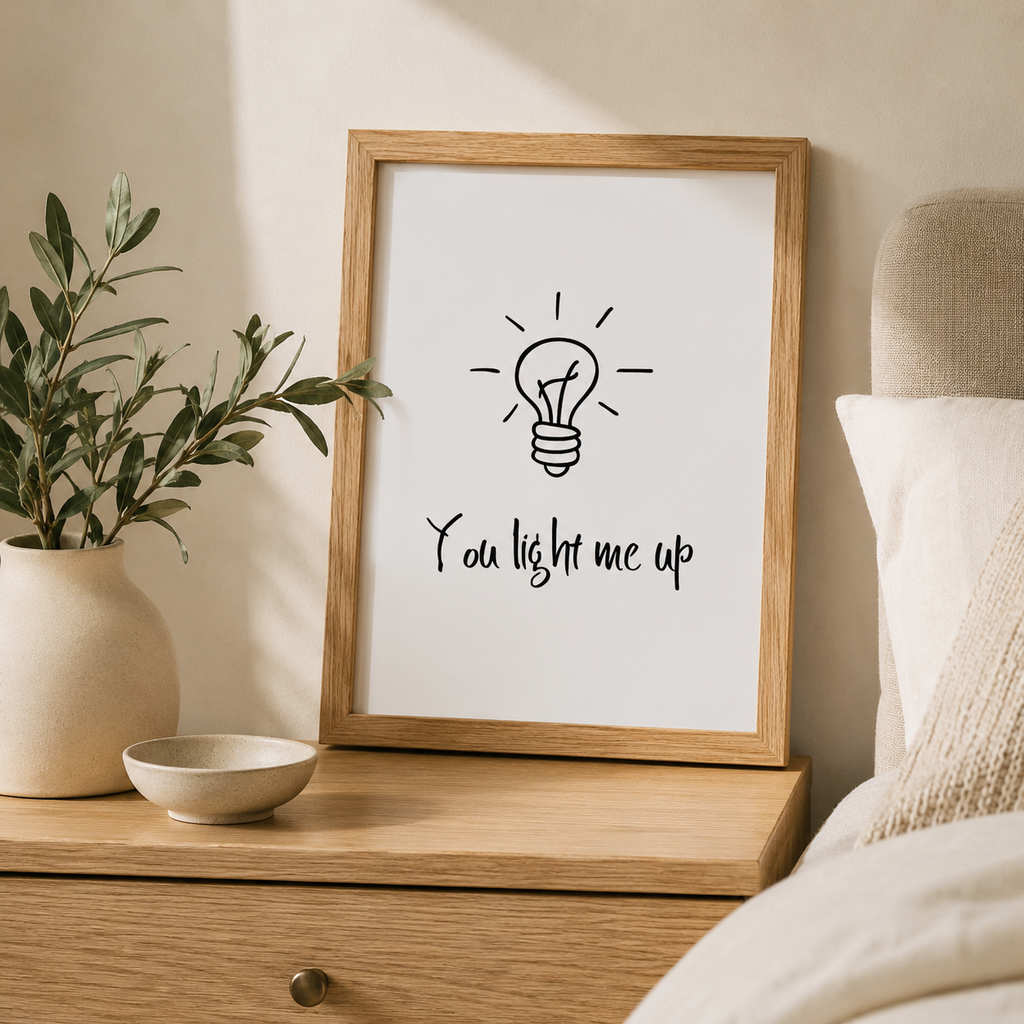 'You light me up' print with a hand-drawn light bulb in an oak frame on a bedside table