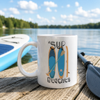 White mug reading 'Sup Beaches' with a paddleboards and paddle design, by a lake