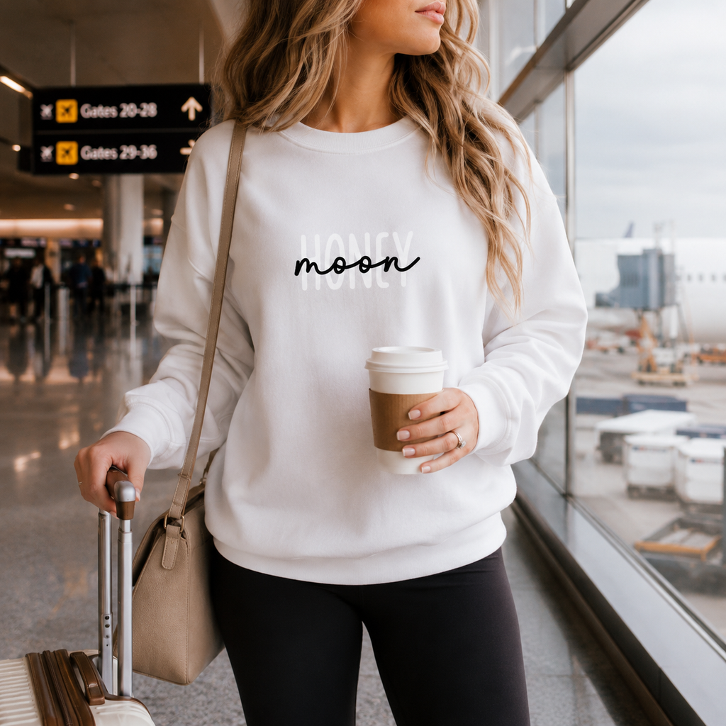 White honeymoon sweatshirt with 'Honeymoon' script, worn by a woman at an airport
