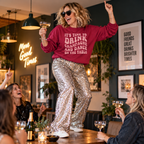Woman wearing the burgundy slogan sweatshirt and sequin trousers, dancing on a table at a party with a champagne glass raised.