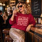 Woman in heart sunglasses wearing the burgundy champagne slogan sweatshirt, holding a glass of champagne at a bar.