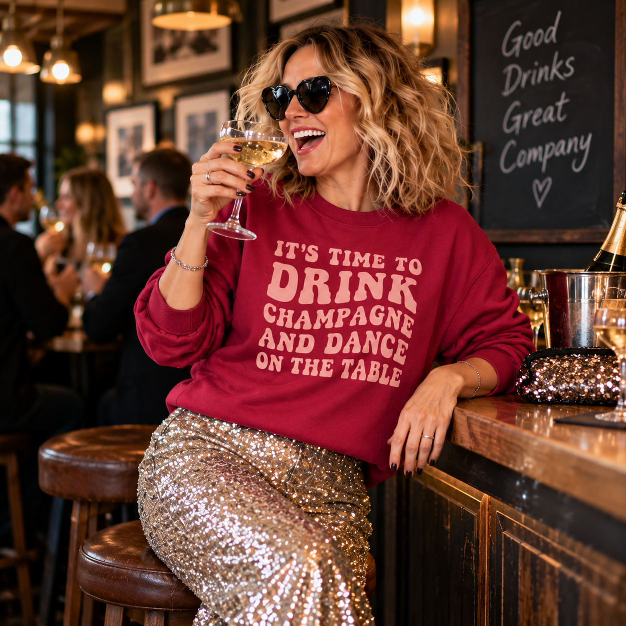 Woman in heart sunglasses wearing the burgundy champagne slogan sweatshirt, holding a glass of champagne at a bar.