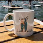 White mug with paddle design and text on a wooden surface by a lake with paddleboarders.