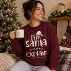 A woman sitting in a cozy holiday setting wearing a maroon crewneck sweatshirt with the white text 'DEAR Santa i can EXPLAIN' and a small Santa hat graphic. She is holding a white mug with a red heart, with a decorated Christmas tree and fireplace in the background.