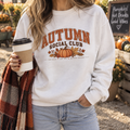 Woman wearing a white Autumn Social Club pumpkin sweatshirt, holding a coffee cup at an autumn pumpkin market stall.