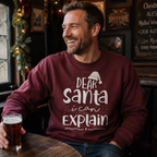 A man in a pub wearing a maroon sweatshirt with the white text 'DEAR Santa i can EXPLAIN' and a small Santa hat graphic. He is holding a pint of beer and smiling while looking to the side.