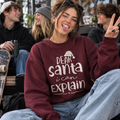 A woman sitting outdoors wearing a maroon crewneck sweatshirt with the white text 'DEAR santa i can EXPLAIN' and a Santa hat graphic. She is making a peace sign with her hand, with other students visible in the background.