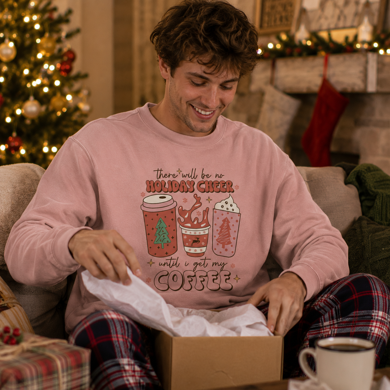 A man sitting on a couch in a cozy living room, opening a box of gift wrapping paper. He is wearing a light pink crewneck sweatshirt with a Christmas-themed coffee graphic and text that reads 'there'll be no HOLIDAY CHEER + until i get my COFFEE', paired with red and black plaid pajama pants.