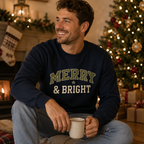 A man wearing a navy blue crewneck sweatshirt with 'MERRY & BRIGHT' printed on the front, sitting in a cozy living room decorated for Christmas.