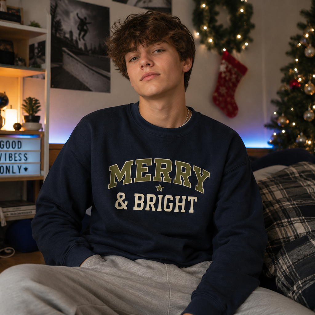 A young man wearing a navy blue crewneck sweatshirt with 'MERRY & BRIGHT' printed in olive green and cream collegiate lettering, sitting in a cozy, decorated room with Christmas lights and a stocking in the background.