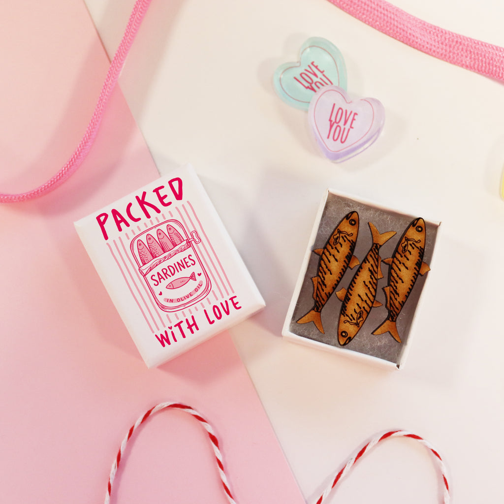 Matchbox with 'Packed with Love' text and illustration of sardines, surrounded by decorative ribbons on a pink background.