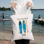 Person holding a tote bag with a paddleboard design and text by a lakeside.