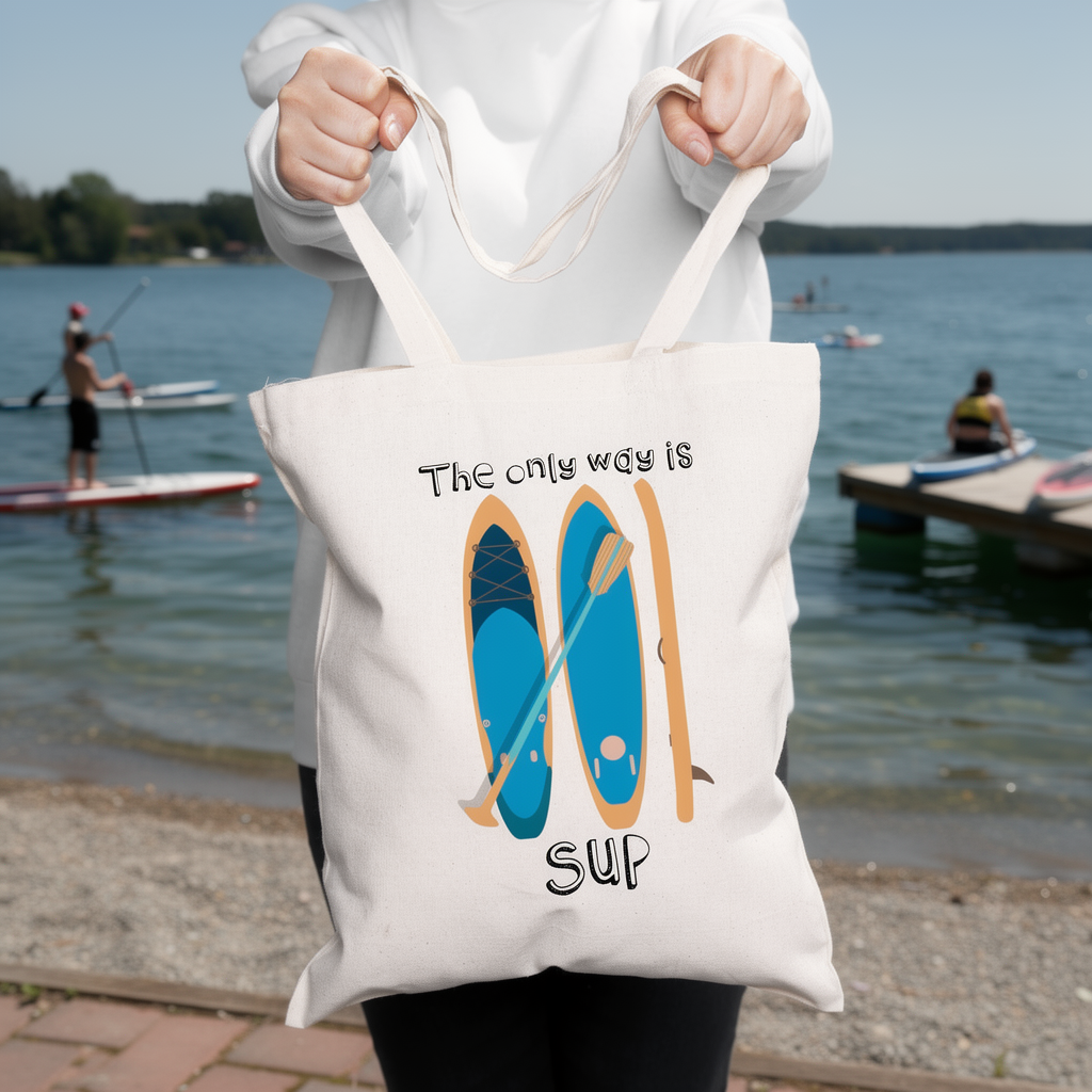 Person holding a tote bag with a paddleboard design and text by a lakeside.