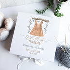 A personalized white luxury baby gift box with hand-drawn illustration, a watercolour bear design, and the name 'Charlotte it's a girl' written on it, alongside some knitting yarn and scissors.