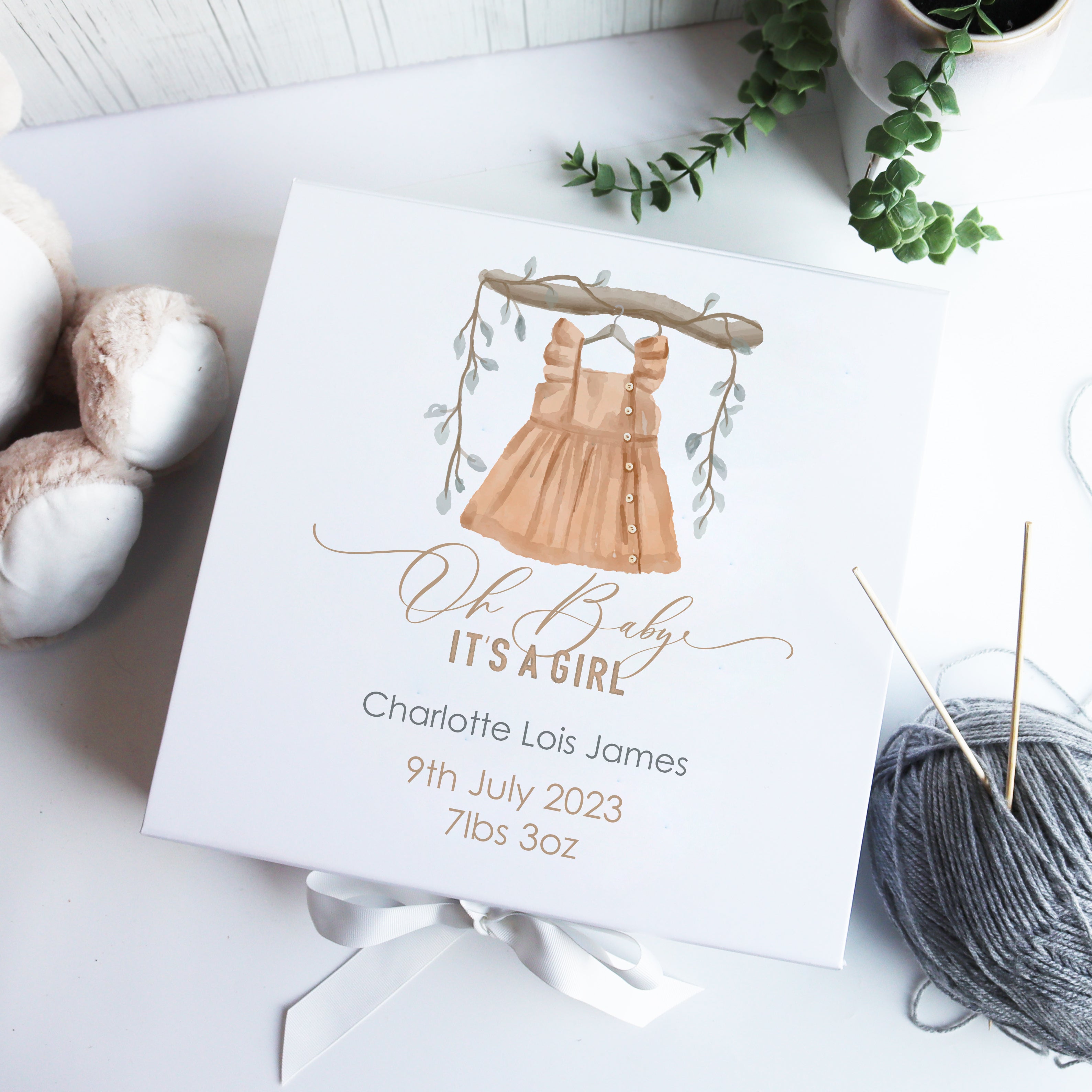 A personalized white luxury baby gift box with hand-drawn illustration, a watercolour bear design, and the name 'Charlotte it's a girl' written on it, alongside some knitting yarn and scissors.