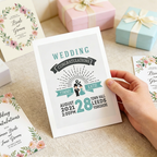 Hand holding a wedding invitation card with details on a table with gift boxes.