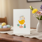 Happy Easter card with a yellow chick design on a table with Easter eggs and flowers.