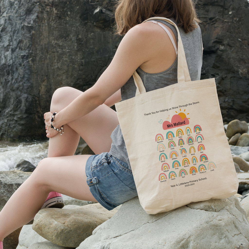 A person sitting by a body of water with a large rock in the foreground, holding a beige tote bag with a colorful rainbow and hearts design and a message that reads 'Thank you for helping us shine through the storm.'.