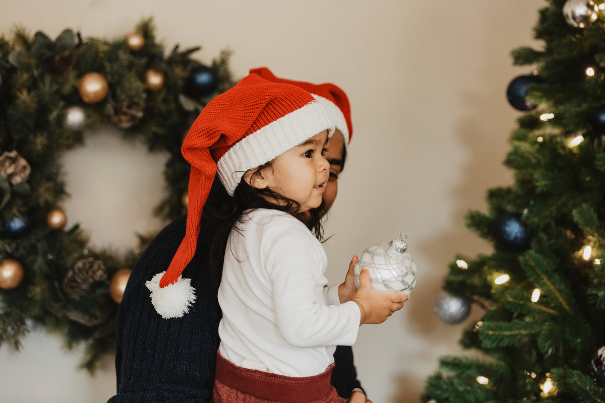 Child in a Santa hat holding a bauble by a Christmas tree