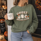 Woman wearing a dusty green Ghost Social Club sweatshirt with the front print, holding a 'Spooky Season' mug on a porch with pumpkins and a skeleton.