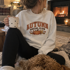 Woman wearing a white Autumn Social Club sweatshirt sitting by a fireside with a dog, holding a heart-patterned mug.