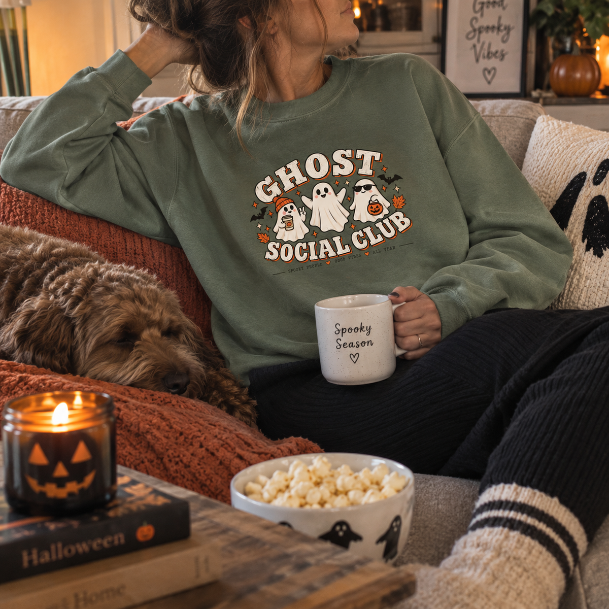 Woman wearing a dusty green Ghost Social Club sweatshirt with the front print, sitting on a sofa with a dog, holding a 'Spooky Season' mug.