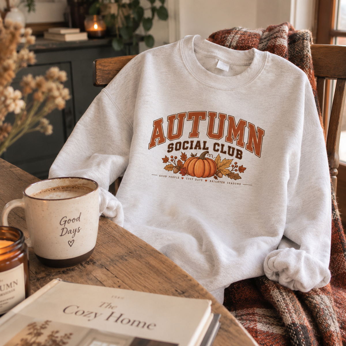 Woman wearing a white Autumn Social Club sweatshirt at a table with a coffee mug, candle and cosy home book.