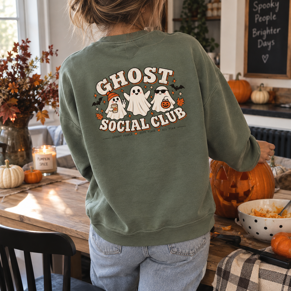 Woman wearing a dusty green Ghost Social Club sweatshirt with the back print, standing in a kitchen with pumpkins and a Halloween chalkboard sign.
