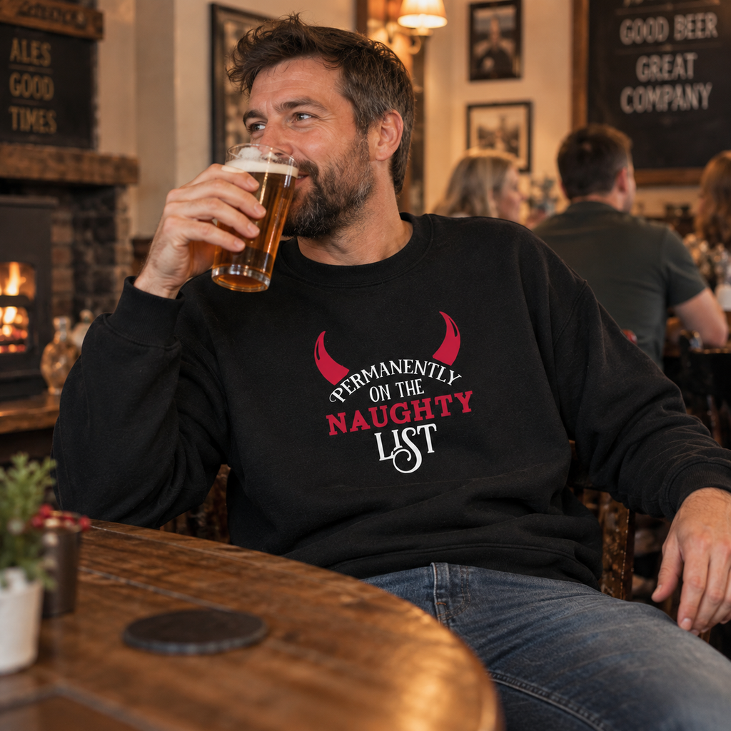 Man wearing a black Permanently On The Naughty List Christmas jumper, holding a pint in a pub.