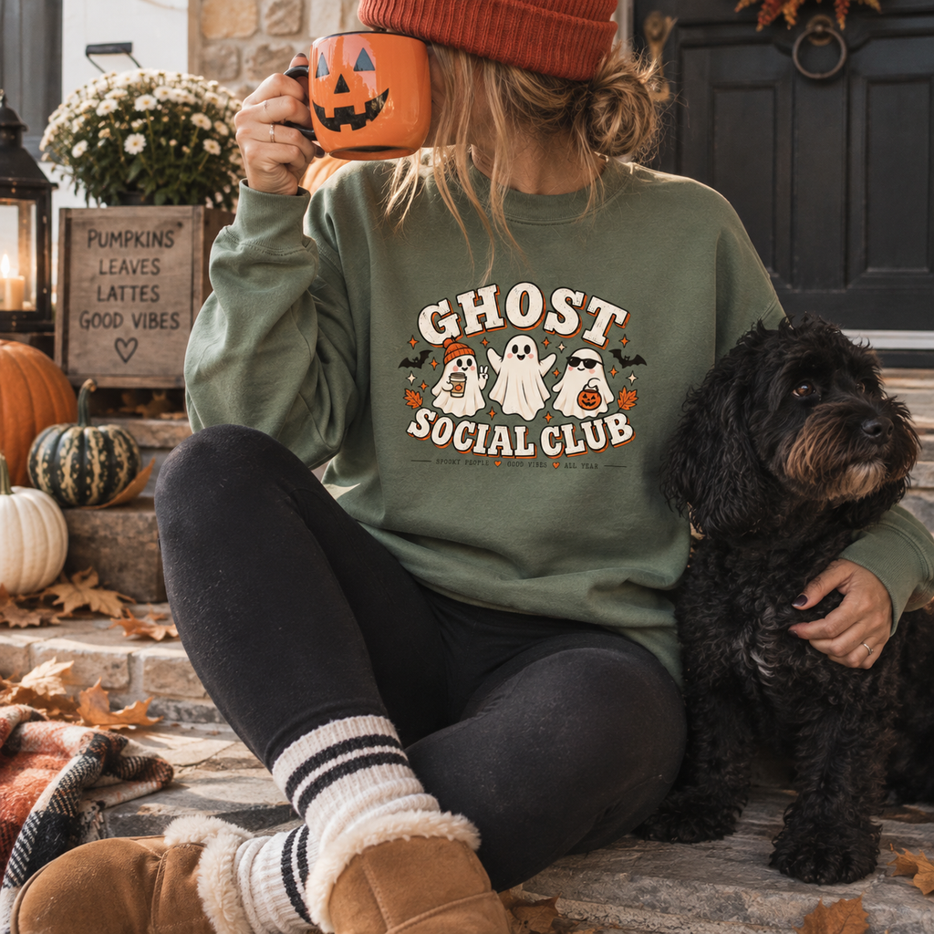 Woman wearing a dusty green Ghost Social Club sweatshirt with the front print, sitting on porch steps with a dog, holding a pumpkin-shaped mug.