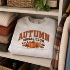 Folded white Autumn Social Club pumpkin sweatshirt on a shelf among knitwear, plants and a plaid throw.