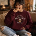 Woman wearing a burgundy Official Mince Pie Tester Christmas jumper, eating a mince pie by a lit fireplace.