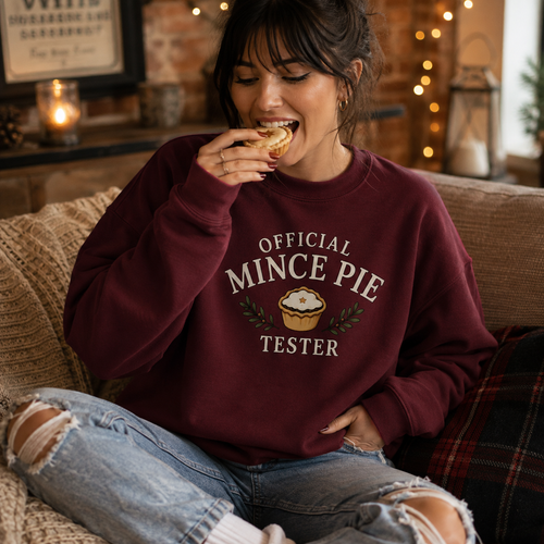 Woman wearing a burgundy Official Mince Pie Tester Christmas jumper, eating a mince pie by a lit fireplace.
