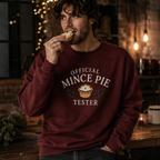 Man wearing a burgundy Official Mince Pie Tester Christmas sweatshirt, eating a mince pie in a festively lit kitchen.