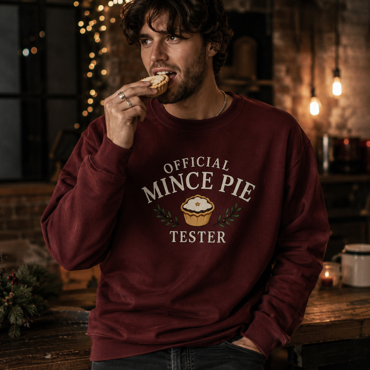 Man wearing a burgundy Official Mince Pie Tester Christmas sweatshirt, eating a mince pie in a festively lit kitchen.