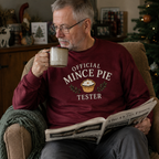 Older man wearing a burgundy Official Mince Pie Tester Christmas jumper, drinking from a mug and reading a newspaper.