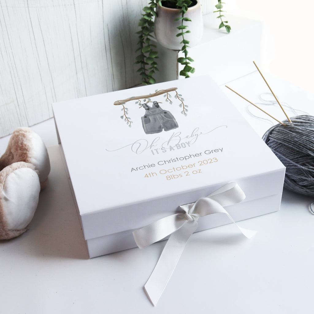 A white luxury baby gift box with a hand-drawn illustration, personalized with a baby boy's name and birth details, adorned with a white ribbon and placed on a wooden surface with a plant in the background.
