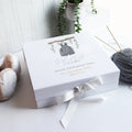 A white luxury baby gift box with a hand-drawn illustration, personalized with a baby boy's name and birth details, adorned with a white ribbon and placed on a wooden surface with a plant in the background.