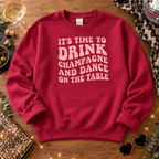 Burgundy champagne and dance slogan sweatshirt laid flat on a wooden table with a disco ball, fairy lights and a glass of champagne.