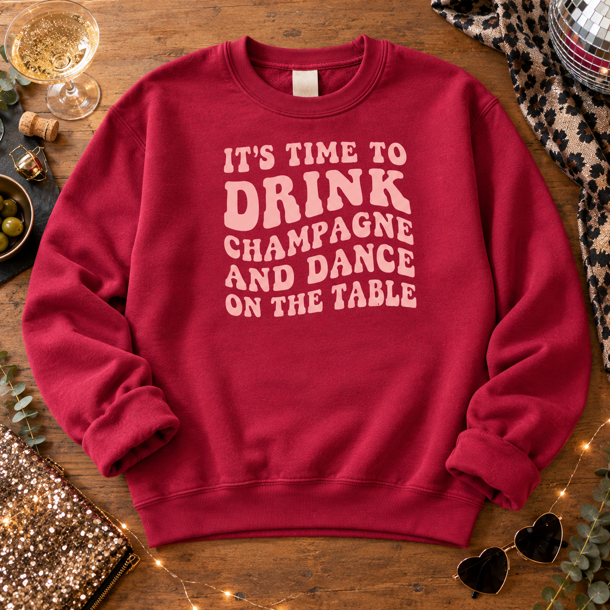 Burgundy champagne and dance slogan sweatshirt laid flat on a wooden table with a disco ball, fairy lights and a glass of champagne.