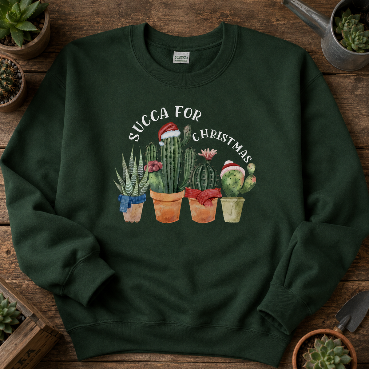 Folded green Succa For Christmas jumper on a wooden table with potted succulents.