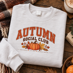 Folded white Autumn Social Club pumpkin sweatshirt on a wooden table with a plaid blanket, coffee and a pumpkin.
