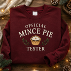 Folded burgundy Official Mince Pie Tester Christmas jumper on a wooden surface with pine sprigs and pinecones.